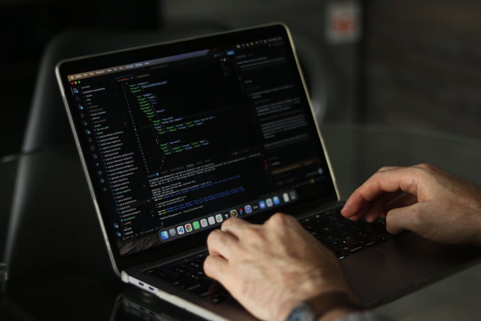A person typing on a laptop with a dark screen displaying lines of code, seated at a black table. The individual's hands are visible, with one hand on the keyboard and the other poised above the track